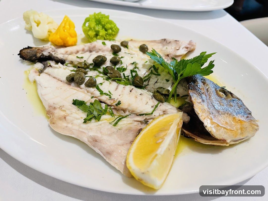 A white plate holds a whole cooked fish, filleted and topped with capers and chopped parsley. A lemon wedge is placed on the side. Accompanying the fish are portions of cauliflower, a yellow puree, and Romanesco broccoli. The dish is presented on a white tablecloth.