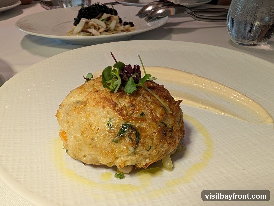 A close-up shot of a crab cake on a white plate, garnished with microgreens and served with a swirl of white sauce and a drizzle of olive oil. In the background, another plate holds a dish with squid and black rice.
