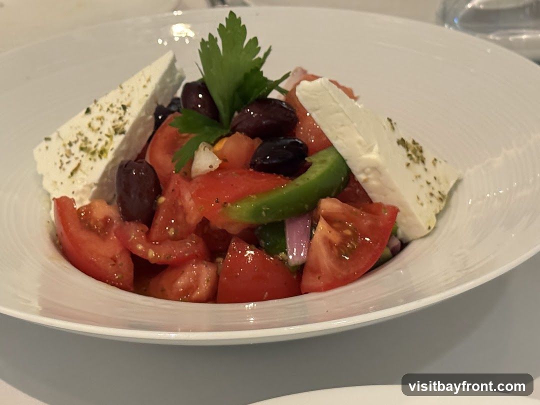 A close-up shot of a Greek salad served in a white bowl. The salad contains chopped tomatoes, Kalamata olives, green bell peppers, red onion, and feta cheese. A sprig of parsley garnishes the top.