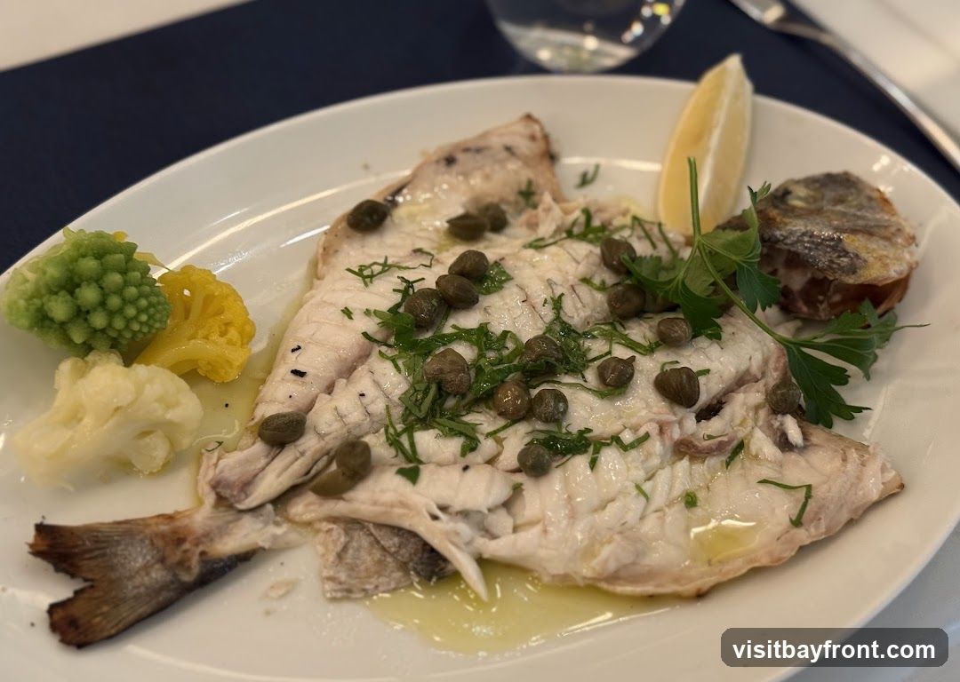 A white oval plate holds a whole cooked fish, garnished with capers, chopped herbs, and a lemon wedge. The fish is served with a side of steamed vegetables, including cauliflower, yellow cauliflower, and Romanesco broccoli. A piece of fried potato is also visible on the side.