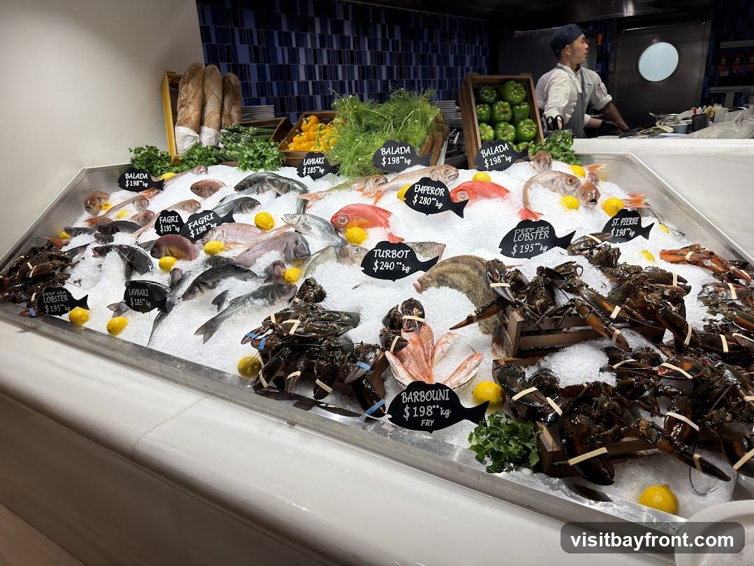 A display of fresh seafood on ice, including lobsters and various types of fish. Each type of seafood has a small black sign with its name and price per kilogram. In the background, a chef is visible working in a kitchen area.