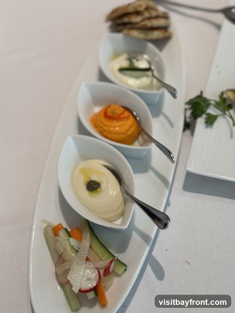 A platter with three small bowls of dips, a side of chopped vegetables, and a stack of pita bread. The dips appear to be hummus, a red pepper dip, and a white yogurt-based dip with cucumber.