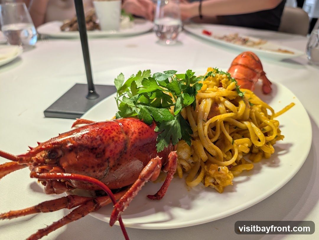 A whole cooked lobster is served on a white plate with a pile of pasta and garnished with parsley. The dish is set on a white tablecloth, with other diners and plates visible in the blurred background.
