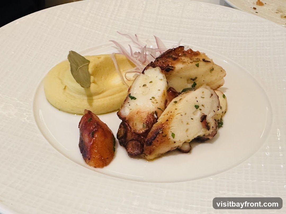 A white plate holds a serving of grilled octopus tentacles, a dollop of yellow puree, and a small piece of roasted potato. The octopus is seasoned with herbs and has grill marks. Thinly sliced red onions are visible behind the puree, and a bay leaf garnishes the puree.