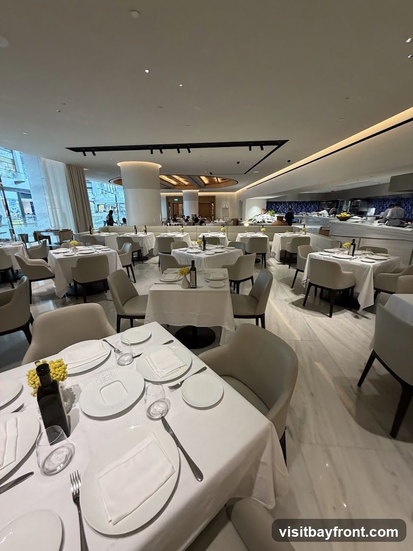 A well-lit, modern restaurant dining room with white tablecloths on multiple tables. Chairs are upholstered in a neutral tone. In the background, an open kitchen area is visible with chefs working. Natural light streams in from large windows on the left.