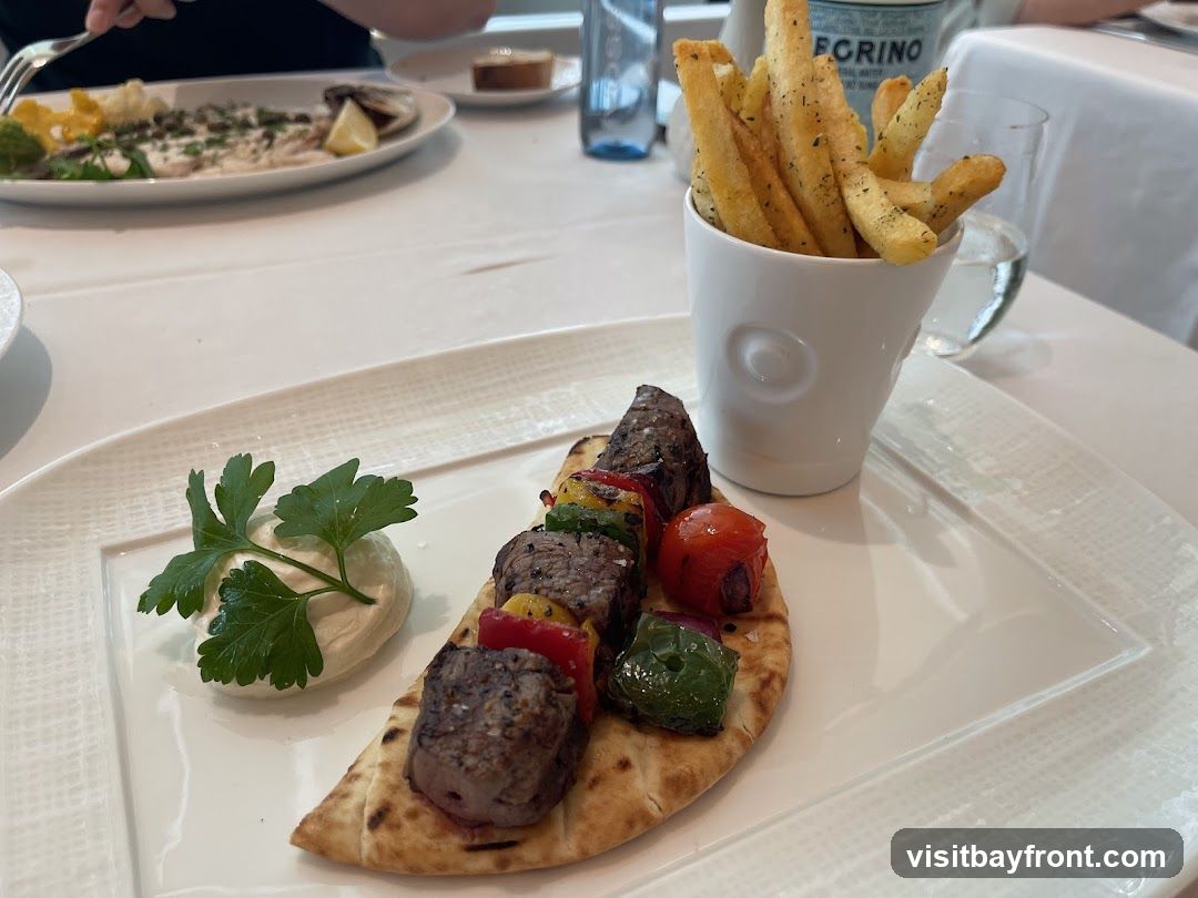 A plate of beef skewers served on pita bread with a dollop of dip and a garnish of parsley. A white cup filled with french fries is placed next to the skewers. In the background, another plate with a fish dish and a glass of water are visible.