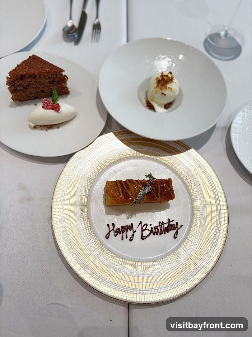A close-up shot of a table setting with three dessert plates. The plate in the foreground has a rectangular pastry topped with herbs and drizzled with caramel, with "Happy Birthday" written in chocolate below it. To the left, a slice of dark cake sits next to a quenelle of ice cream garnished with a raspberry and mint leaf. To the right, a white bowl contains a scoop of ice cream with a sprinkle of nuts and caramel sauce.