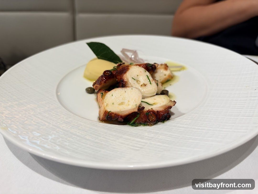 A white plate holds a serving of grilled octopus. The octopus is cut into pieces and garnished with herbs and capers. A dollop of yellow puree, possibly mashed potatoes or polenta, is also on the plate. A person's shoulder and arm are visible in the blurred background.