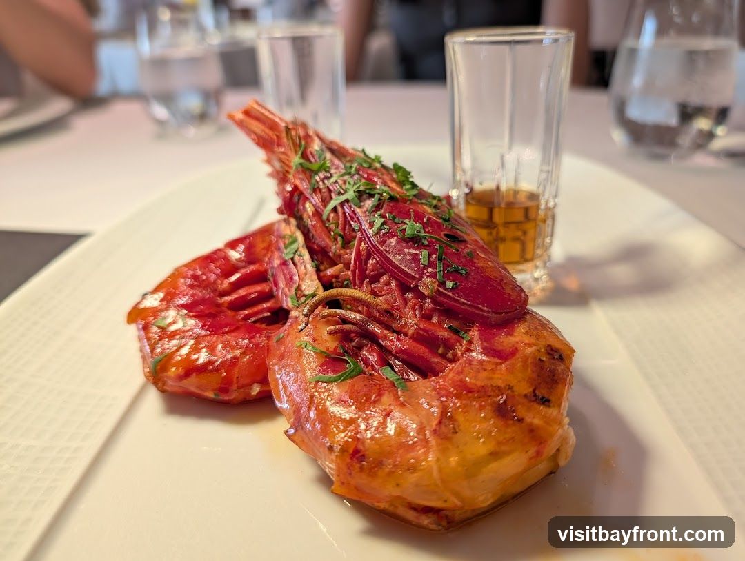 A close-up shot of a gourmet dish featuring three large, red prawns, garnished with chopped herbs. The prawns are served on a white, textured rectangular plate. In the background, out of focus, are glasses of water and a small shot glass containing amber liquid, suggesting a dining setting.