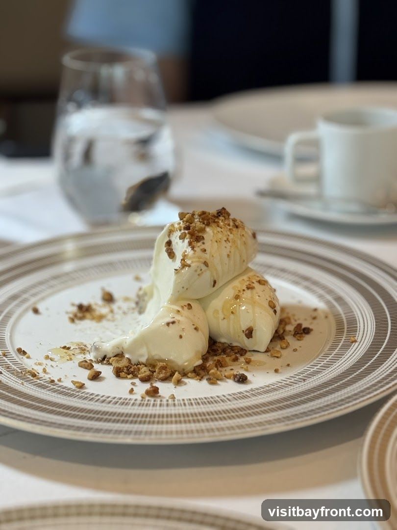 A close-up shot of a dessert, likely ice cream or gelato, served on a decorative plate. The dessert is topped with chopped nuts and drizzled with honey. A glass of water and a cup of coffee are visible in the blurred background, suggesting a dining setting.