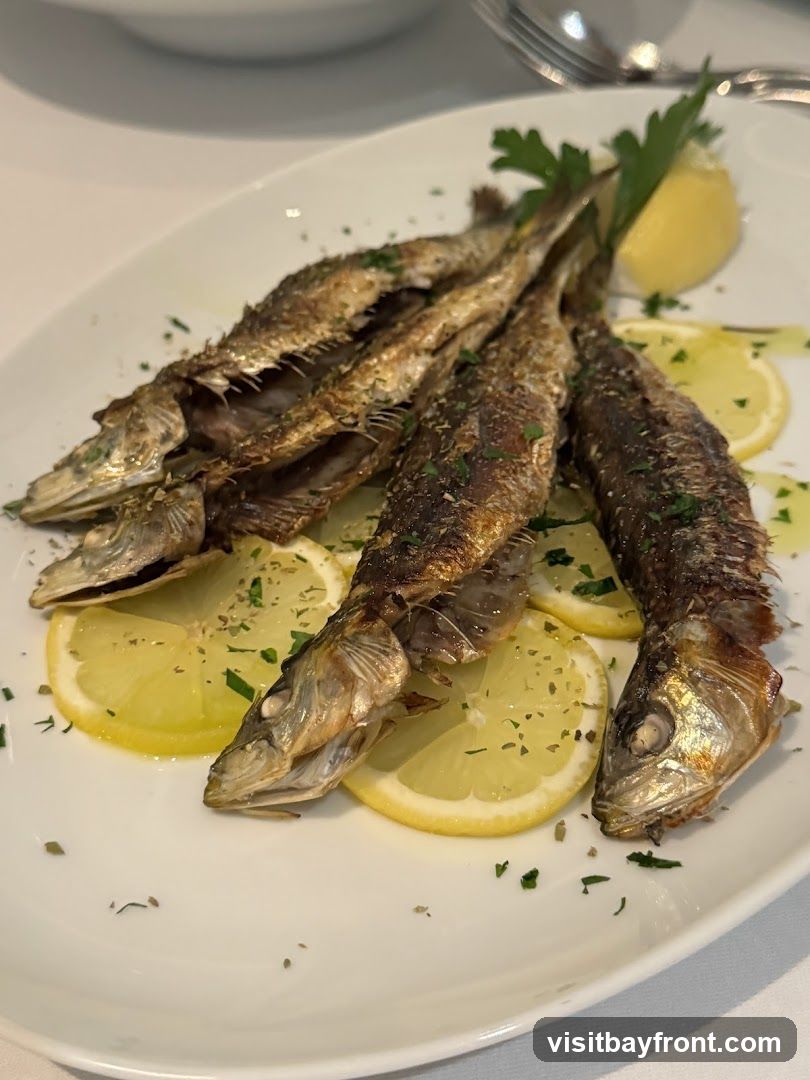 A white oval platter holds several small, whole fried fish arranged on top of lemon slices. The fish are golden brown and appear to be seasoned with herbs. A sprig of parsley garnishes the dish. The platter is set on a white tablecloth.