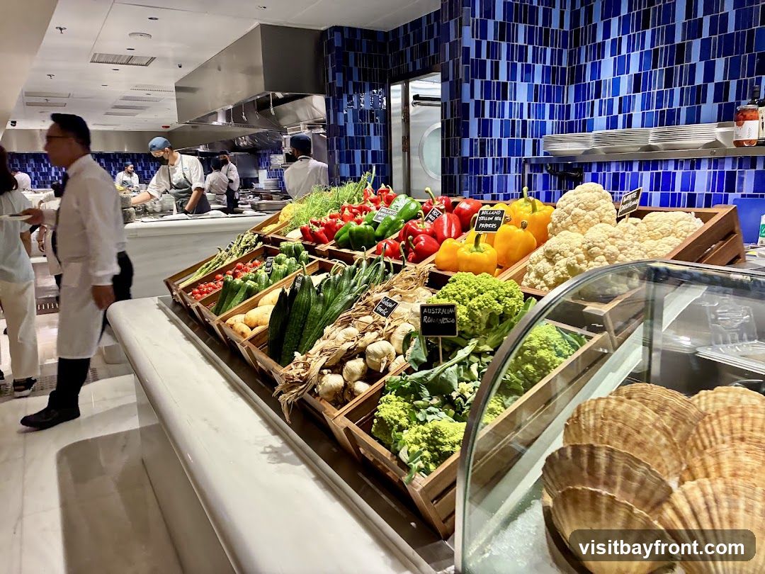 A buffet display featuring a variety of fresh vegetables and seafood. In the foreground, large scallops are visible in a refrigerated case. Behind the scallops, wooden crates are filled with produce including cauliflower, romanesco broccoli, bell peppers, garlic, and various leafy greens and root vegetables. In the background, chefs are working in a kitchen area with stainless steel equipment and blue tiled walls.