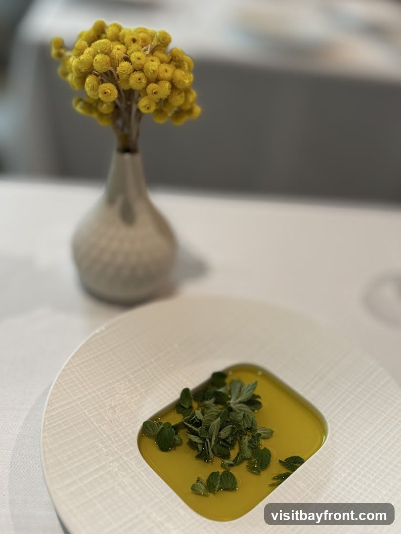 A close-up shot of a white, textured plate holding a square of golden liquid, possibly oil or broth, topped with fresh green herbs. In the blurred background, a small, light-colored vase holds a cluster of yellow, dried flowers. The setting appears to be a restaurant table with white tablecloths.