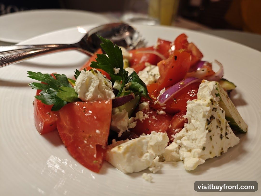 A close-up shot of a Greek salad on a white plate. The salad contains chopped tomatoes, red onion, cucumber, feta cheese, and parsley. A fork and spoon are visible in the background.