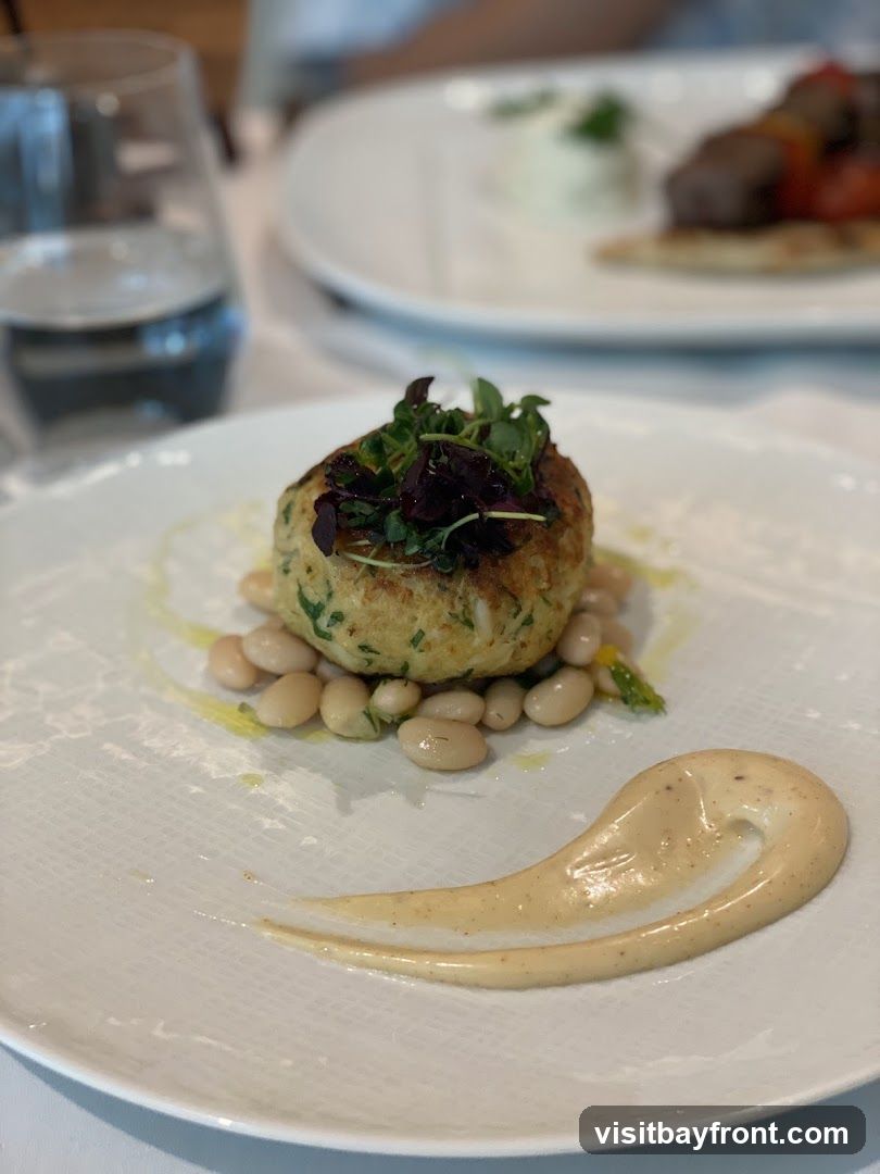 A close-up shot of a crab cake served on a white plate with white beans and a swirl of sauce. The crab cake is topped with microgreens. In the background, another plate with food and a glass of water are visible.