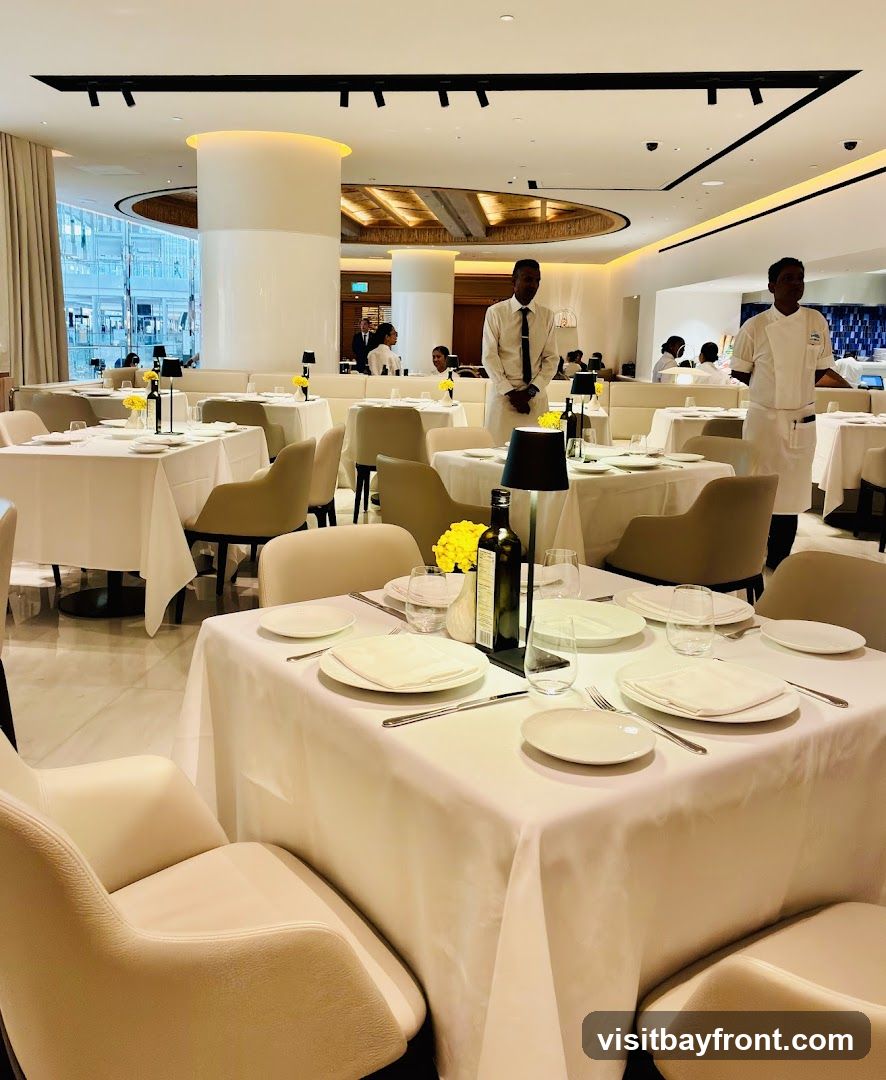 A well-lit, upscale restaurant interior with white tablecloths on multiple tables. Staff members in uniform are visible, attending to the dining area. The decor is modern and minimalist, with large windows offering a view of the outside.