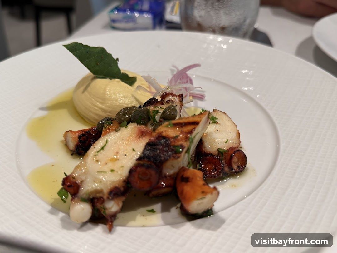 A close-up shot of a grilled octopus appetizer served on a white textured plate. The dish features several pieces of grilled octopus tentacles, garnished with capers, red onion slivers, and fresh herbs. A dollop of light yellow puree, possibly hummus or a vegetable spread, is placed on the side, topped with a green leaf. The octopus appears tender with char marks from grilling, and it is drizzled with olive oil. The background is blurred, showing a table setting with a glass and other plates.