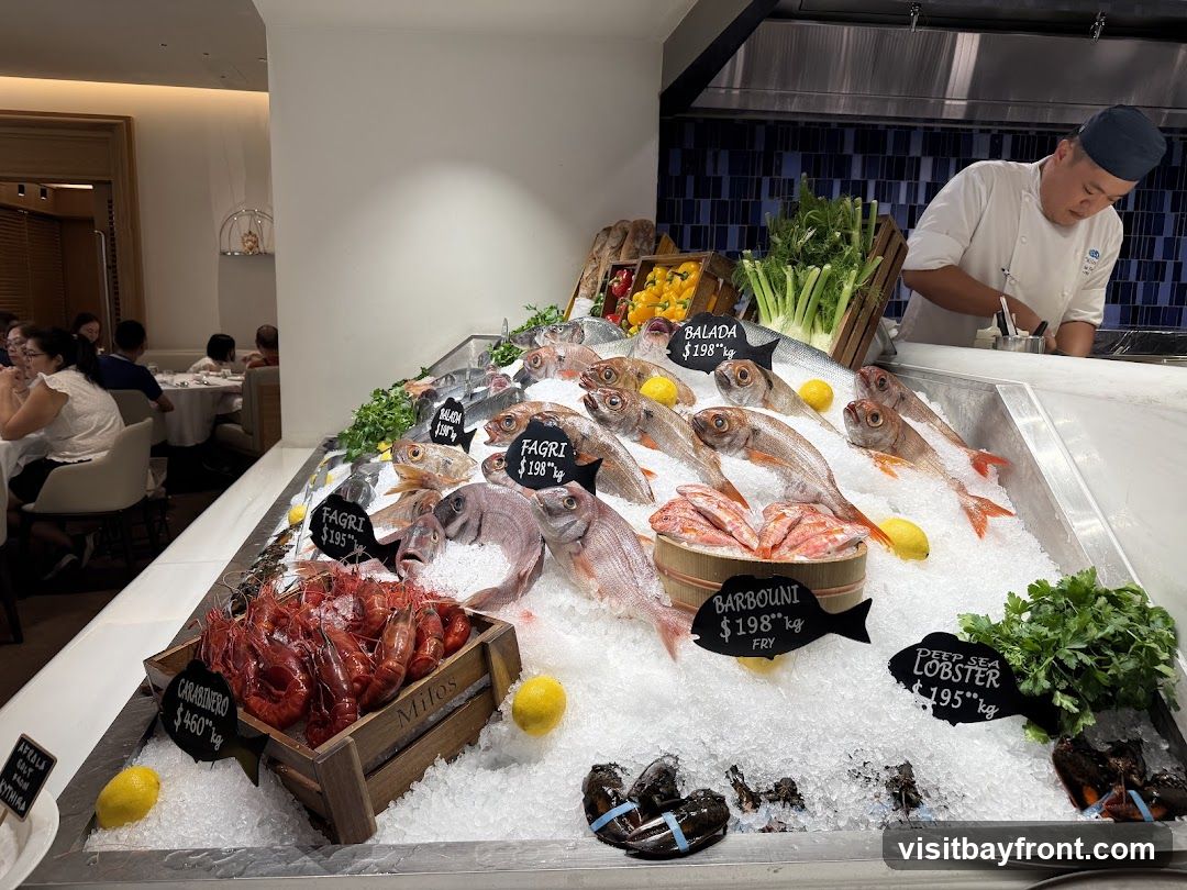 A chef in a white uniform and blue hat prepares seafood behind a display of fresh fish, lobsters, and prawns on ice. Various types of fish are labeled with prices per kilogram, including Balada, FAGRI, and Barbouni. A crate of Carabineros prawns is also visible. In the background, diners are seated at tables in a restaurant setting.