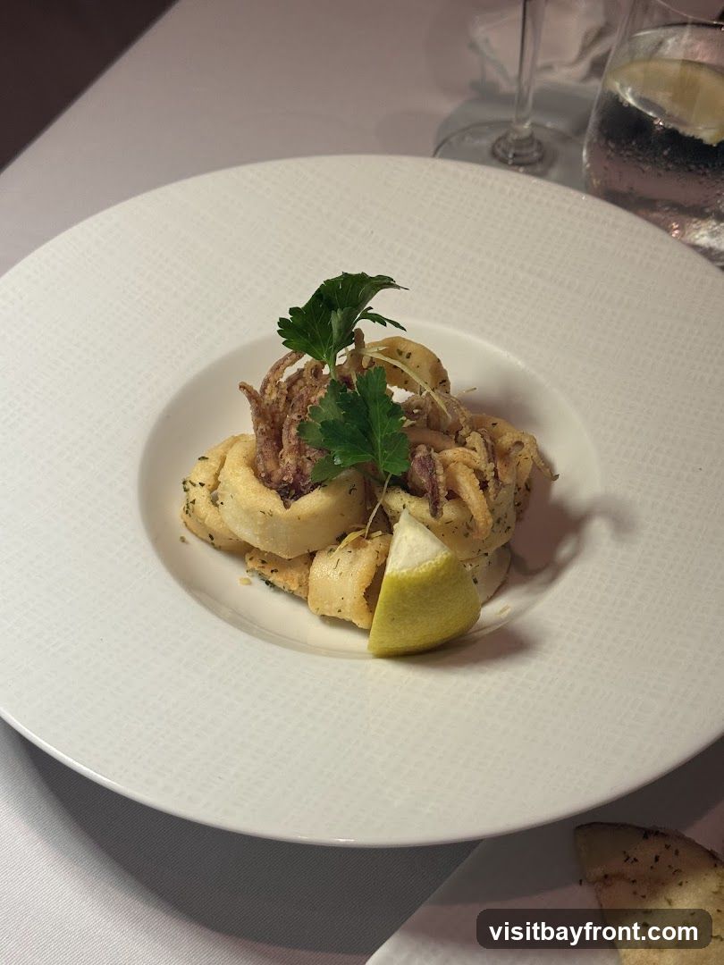 A white plate holds a serving of fried calamari, garnished with parsley and a lemon wedge. The plate is set on a white tablecloth, with a glass of water and a wine glass visible in the background.