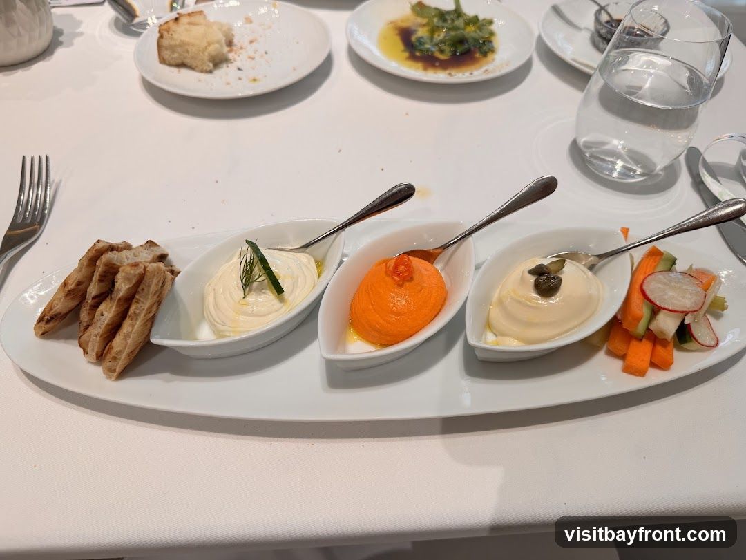 A white platter holds three small dishes of dips, accompanied by breadsticks and crudités. The first dish contains a creamy white dip garnished with dill. The second dish holds a vibrant orange dip. The third dish has a pale yellow dip topped with capers. To the left are several grilled breadsticks, and to the right are sliced carrots, radishes, and cucumber. In the background, other plates with food and a glass of water are visible on a white tablecloth.
