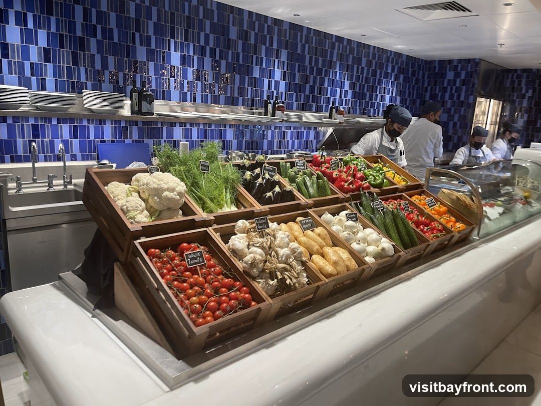 A display of fresh vegetables including cauliflower, fennel, eggplant, zucchini, bell peppers, tomatoes, garlic, and potatoes. Several staff members in chef uniforms are visible in the background, preparing food. The display is set against a backdrop of blue tiled walls.