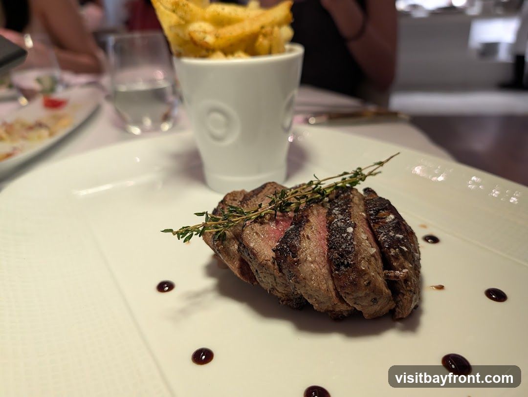 A plate of sliced steak, seasoned with salt and garnished with thyme, is served with a side of french fries in a white ceramic cup. The steak is cooked to a medium-rare. The plate is decorated with small dots of dark sauce. In the background, a glass of water and other diners are visible.