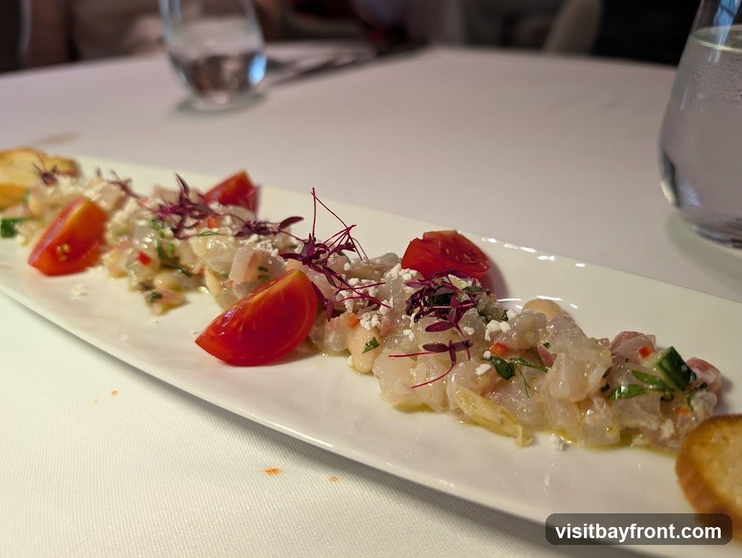 A white rectangular plate holds a serving of ceviche, garnished with chopped tomatoes, microgreens, and crumbled cheese. Toasted bread slices are placed at either end of the plate. The dish is presented on a white tablecloth, with blurred glasses of water in the background.