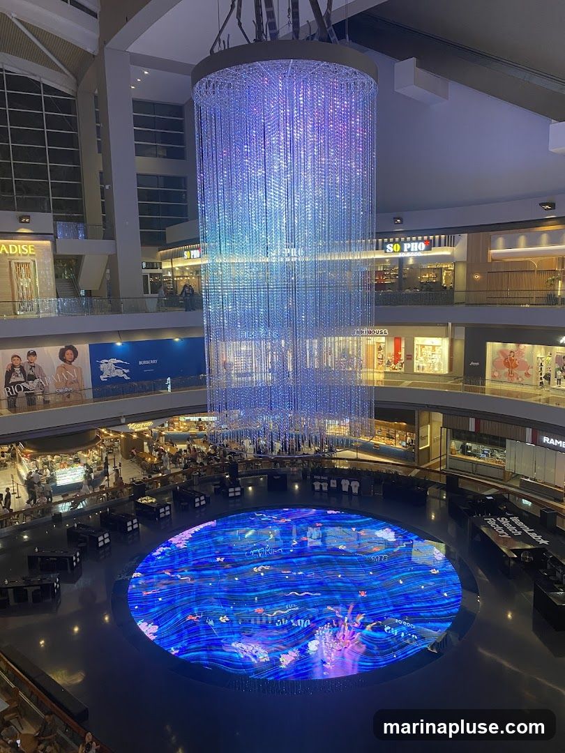An interior view of a multi-level shopping mall atrium. A large, cylindrical chandelier made of numerous strands of lights hangs from the ceiling, casting a blue and purple glow. Below, a circular digital display on the floor shows a vibrant blue aquatic scene with animated elements. Surrounding the display are tables and chairs, suggesting a seating or dining area. Shops and escalators are visible on the upper levels, with signage for brands like "PARADISE", "SO PHO", and "BURBERRY".