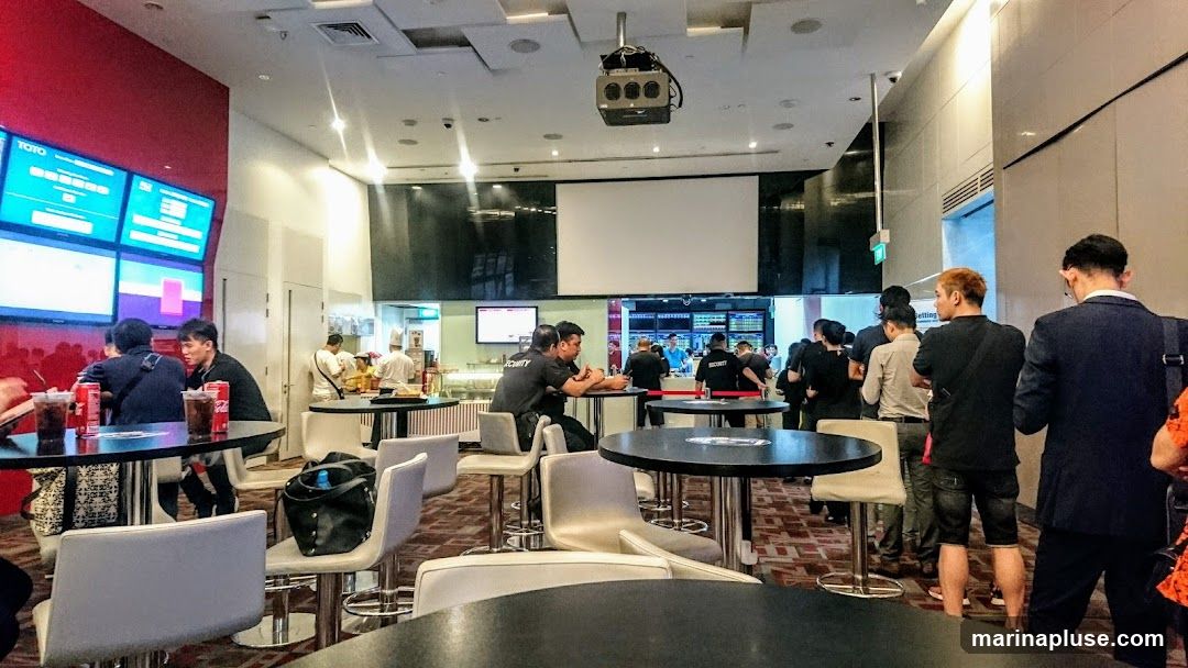A group of people are gathered inside a brightly lit establishment, possibly a betting shop or a sports bar. Several large screens display information, and some individuals are seated at tables with drinks. Others are standing in line or interacting with staff behind a counter. Security personnel are also visible.