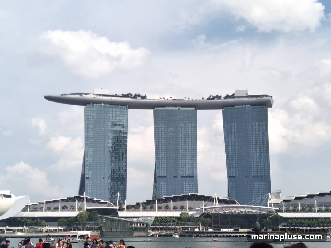 A wide shot of the Marina Bay Sands hotel in Singapore, with its distinctive three towers connected by a sky-high platform. The hotel is set against a cloudy sky. In the foreground, a body of water is visible with a few boats and a crowd of people on a promenade.