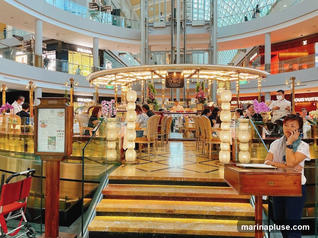 An interior view of a luxurious tea house with ornate lighting and seating. People are dining and conversing. In the background, high-end retail stores like Prada and Louis Vuitton are visible within a modern shopping mall. A menu stand is prominently placed near the entrance.