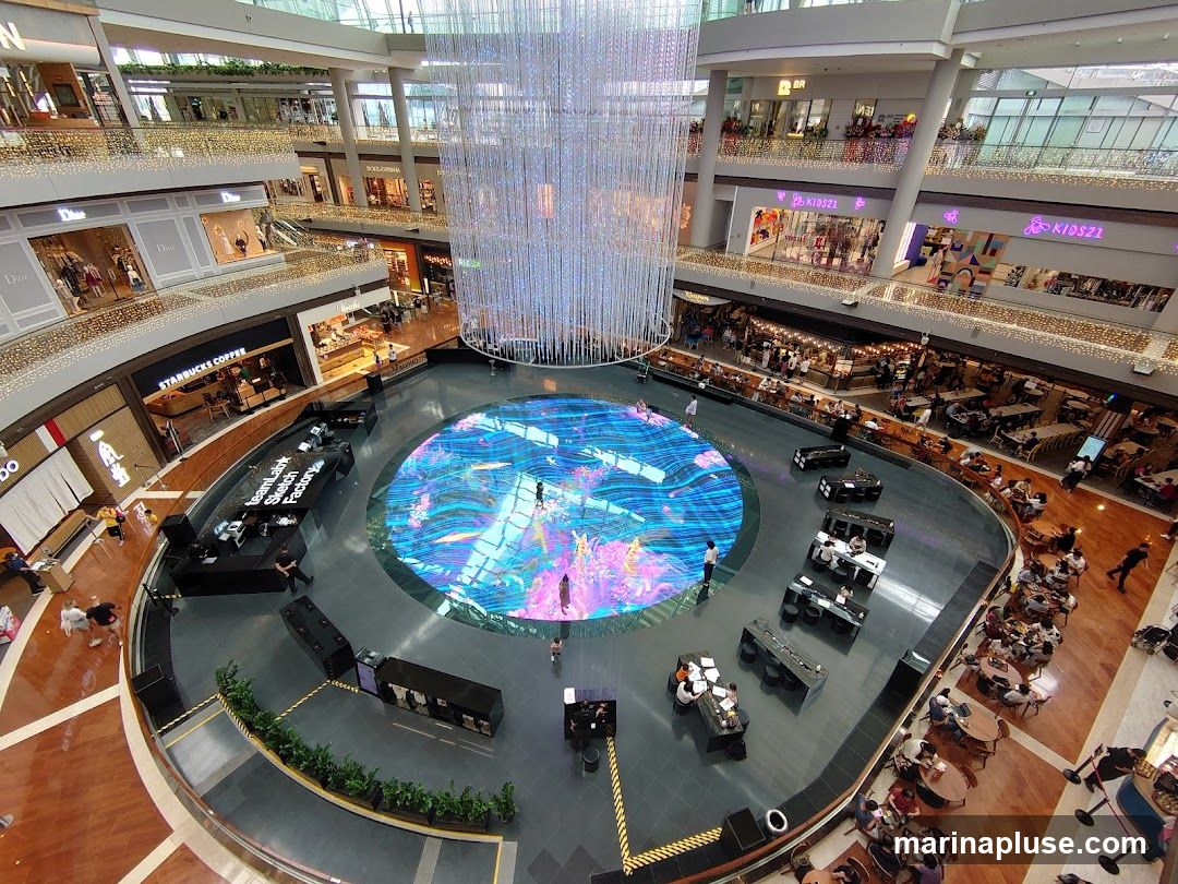 An interior view of a multi-level shopping mall atrium. A large, circular digital display on the floor shows a vibrant, animated aquatic scene. Above the display hangs a massive, shimmering chandelier made of numerous small lights. Several levels of shops and restaurants surround the atrium, with many patrons visible. In the foreground, there are several black kiosks or booths set up around the edge of the digital display.