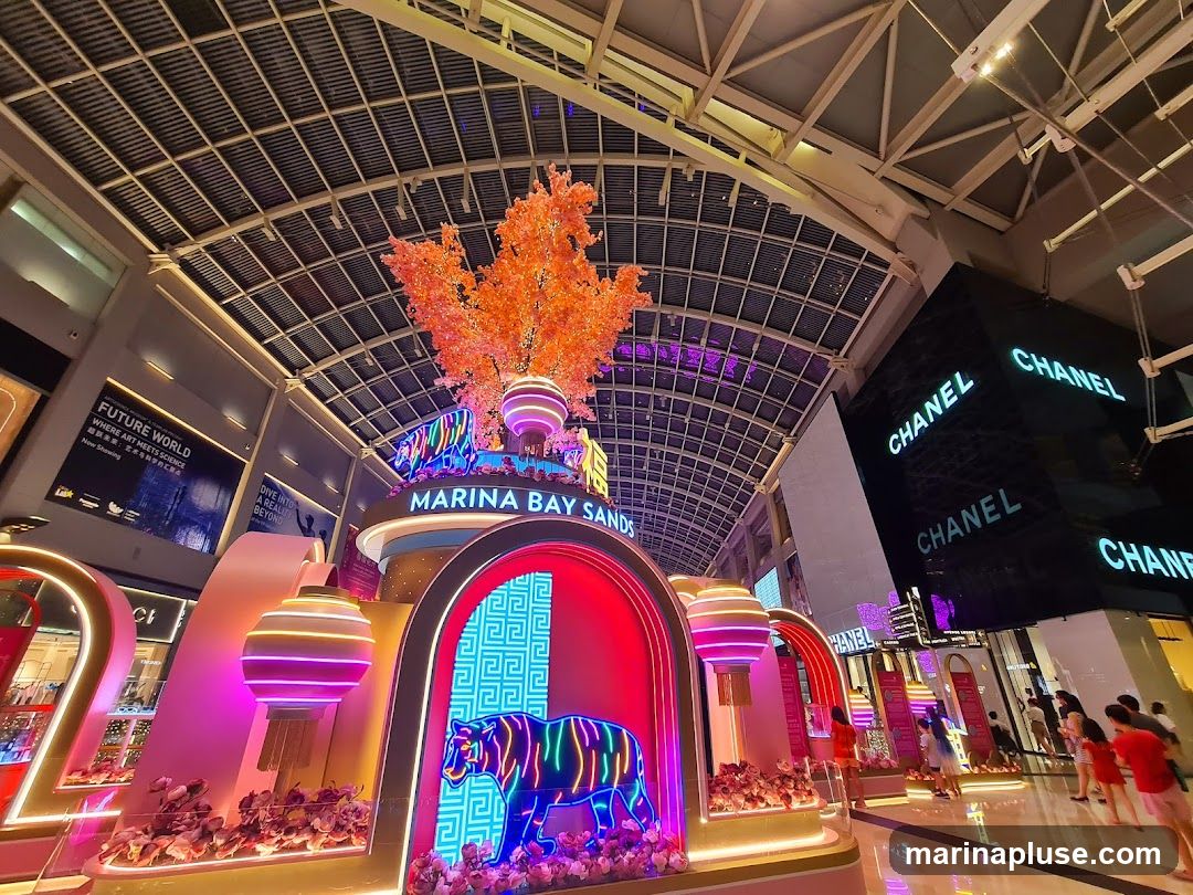 A festive indoor display at Marina Bay Sands, featuring a large, illuminated tiger sculpture with neon accents, surrounded by decorative lanterns and floral arrangements. The display is set against the backdrop of the mall's architecture, including the prominent CHANEL store signage.