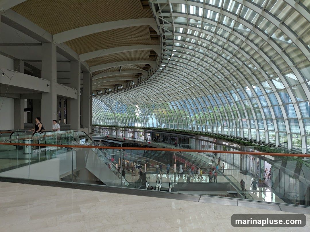 The image shows the interior of a large, modern shopping mall. A distinctive curved glass ceiling arches over the space, allowing natural light to flood in and offering views of the city skyline and water outside. Below the ceiling, multiple levels of the mall are visible, with escalators and walkways connecting them. Shoppers can be seen browsing in stores and moving between levels. The architecture is sleek and contemporary, with polished floors and glass railings.