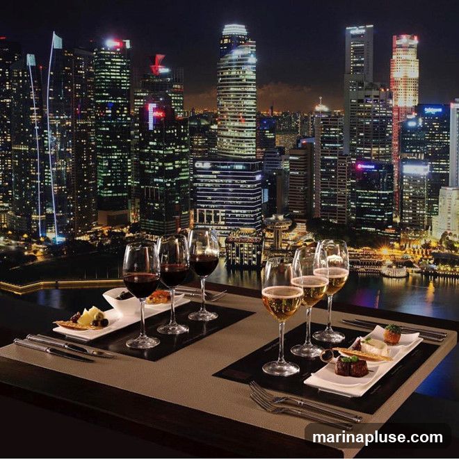 A table set for a meal with wine and appetizers, overlooking a glittering cityscape at night. The table has several glasses of red wine and champagne, along with small plates of food. The background features a dense collection of illuminated skyscrapers.