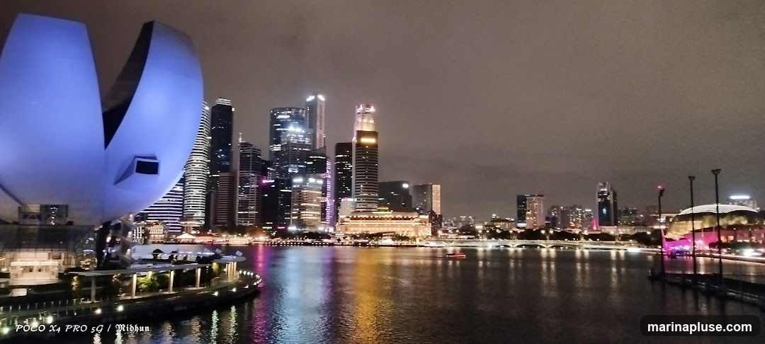 A panoramic view of the Singapore skyline at night, with the Marina Bay Sands resort and other skyscrapers illuminated. The water in the foreground reflects the city lights.