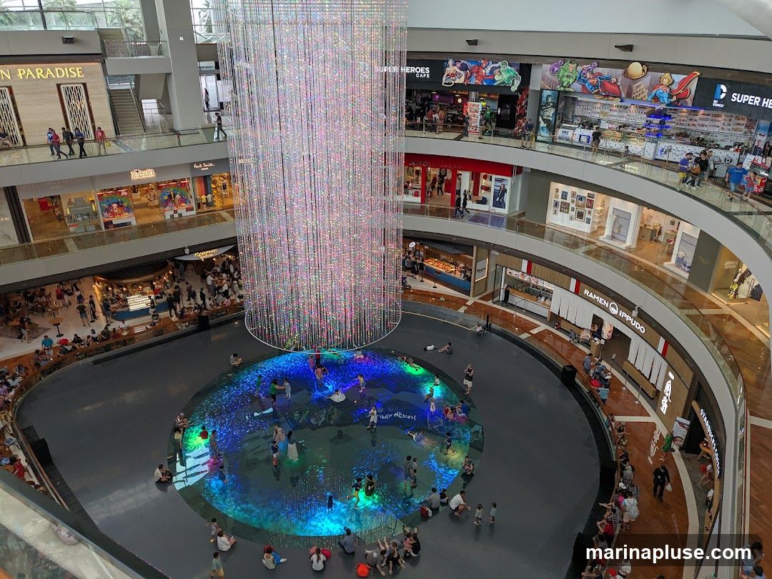 An aerial view of a multi-level indoor shopping mall. In the center is a large, circular water feature with colorful lights and people wading in it. Above the water feature hangs a massive, sparkling crystal chandelier. Surrounding the water feature are various shops and restaurants on different levels, including 'Ramen Ippudo', 'Starbucks Coffee', and a 'Super Heroes Cafe'. People are seen walking around the mall and dining at the establishments.