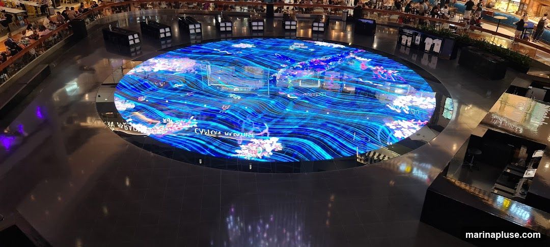 A large, circular digital display is the focal point of a mall atrium. The display shows a vibrant, animated scene of blue waves and colorful, abstract shapes resembling coral or clouds. Text is visible on the display, including what appears to be "CYRILLIC CRUISE" and "BAKEDKA". Surrounding the display is a dark, reflective floor. In the background, there are multiple levels of the mall with shops, seating areas, and people browsing. Some shops have displays of merchandise, including t-shirts.