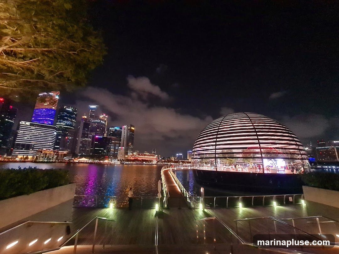 A nighttime view of a unique, domed building with a glass facade, illuminated by interior lights. The building is situated on the water, with a walkway leading to it. In the background, a city skyline with illuminated skyscrapers is visible across the water. The scene is set against a dark night sky with some clouds.