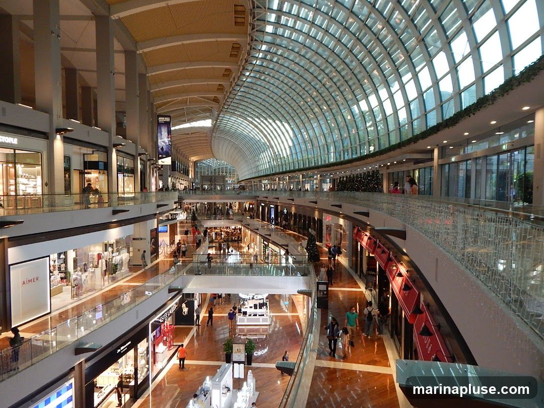 The image shows the interior of a multi-level shopping mall with a distinctive curved glass roof. Shops are visible on multiple floors, with escalators and walkways connecting them. People are seen browsing and walking through the mall. The architecture is sleek and contemporary, with polished floors and modern lighting.