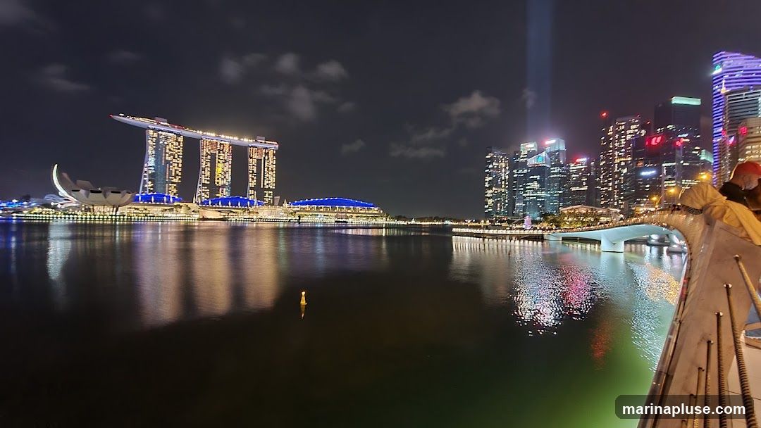 A panoramic view of the Singapore skyline at night, featuring the Marina Bay Sands hotel and other illuminated skyscrapers reflected in the water. A bridge spans the water in the foreground, with a railing and some structures visible on the right side of the image.