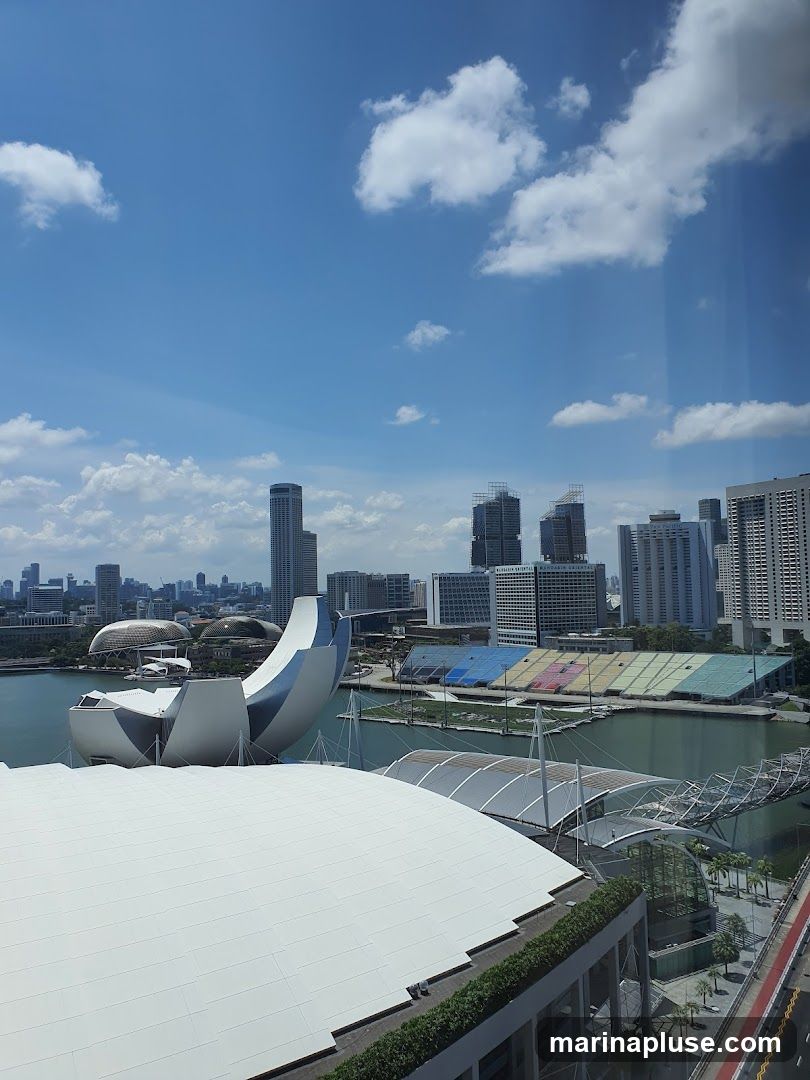 A panoramic view of a modern cityscape under a bright blue sky with scattered white clouds. In the foreground, a large white domed structure is visible, with a sleek, silver, boat-like sculpture in the middle ground. Beyond this, a body of water reflects the sky, and across it, a stadium with colorful seating is apparent. The background is dominated by a dense collection of high-rise buildings, including the distinctive Marina Bay Sands hotel. The overall scene depicts a vibrant urban environment with striking architectural landmarks.