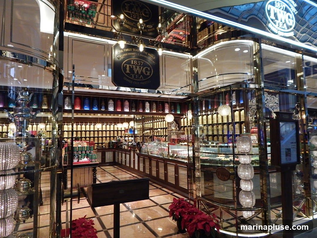 The interior of a TWG Tea shop, featuring shelves stocked with numerous tea canisters of various colors and sizes. Display cases showcase pastries and desserts. Decorative lighting and ornate mirrors adorn the space, creating a luxurious ambiance. A sign with the TWG Tea logo is visible.