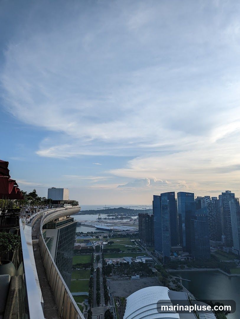 A panoramic view from a high vantage point, likely a rooftop bar or restaurant, overlooking a sprawling city skyline. The foreground features a curved railing and a glimpse of outdoor seating with red umbrellas. In the mid-ground, a modern building with a distinctive curved roof is visible. The background showcases a dense collection of skyscrapers, a body of water, and distant industrial structures under a partly cloudy sky. The lighting suggests it is late afternoon or early evening.