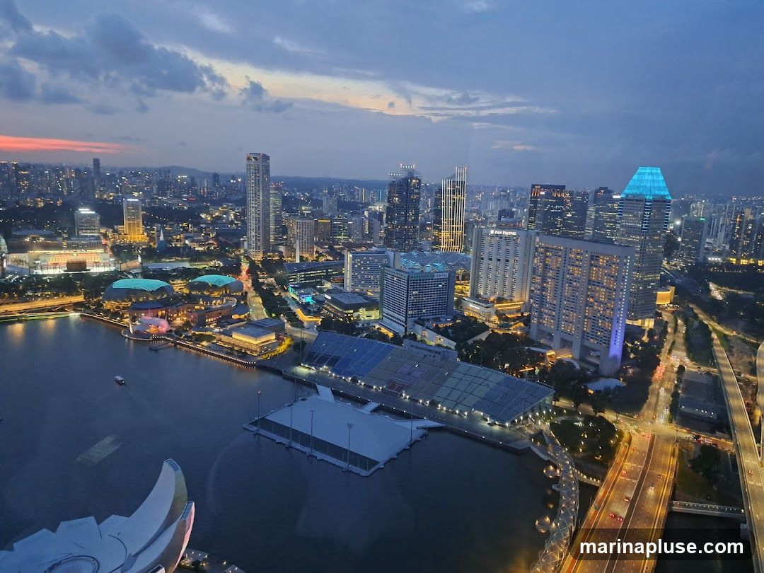 An aerial view of a sprawling cityscape at dusk, with numerous skyscrapers illuminated against a twilight sky. A body of water, likely a bay or river, is in the foreground, reflecting the city lights. Several prominent buildings and structures are visible, including what appears to be a convention center or entertainment complex with distinctive dome-like roofs, and a large hotel with illuminated windows. Highways with moving traffic are also visible, adding to the sense of urban activity. The overall scene captures the vibrant energy of a modern city as day transitions into night.