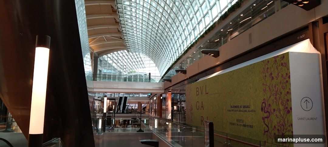 The image shows the interior of a large, modern shopping mall with a distinctive curved glass roof. Escalators and multiple levels of shops are visible. A large banner with floral patterns and text including "BVLGARI", "BUSINESS AS USUAL", and "SAINT LAURENT" is prominently displayed.