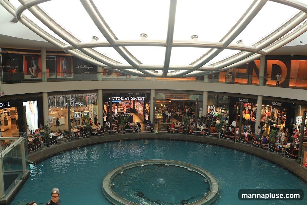 An interior view of a multi-level shopping mall featuring a large indoor fountain. Shops such as Victoria's Secret, Swarovski, and Fendi are visible on the lower levels, with shoppers seated at cafes and browsing the stores. The mall has a modern architectural design with a large, curved glass ceiling.