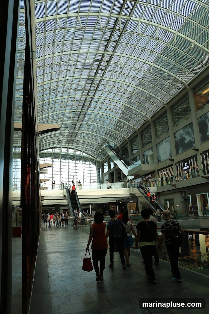 A wide-angle view of the interior of a large, modern shopping mall with a distinctive glass and metal roof structure. People are seen walking along the main walkway, with escalators and multiple levels of shops visible in the background. Some shop signs are discernible, including 'HUBLOT' and 'GAP'. The overall atmosphere is bright and airy, with natural light filtering through the expansive ceiling.