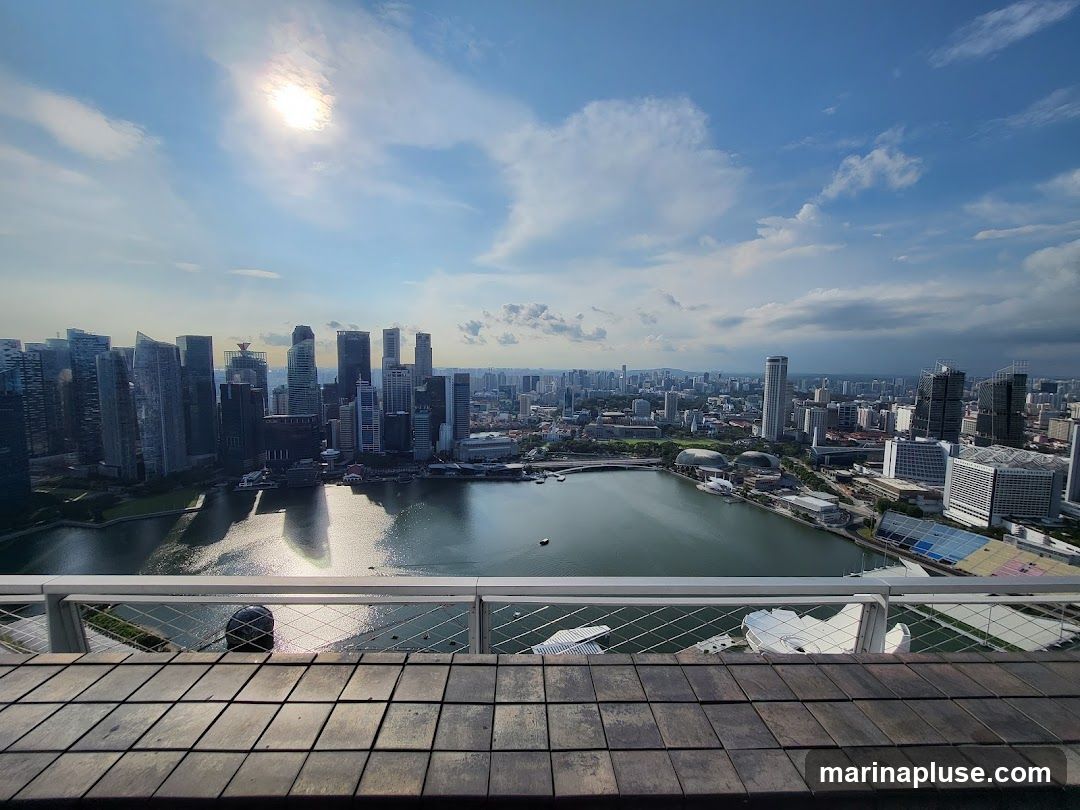 The image captures a sprawling cityscape under a bright, partly cloudy sky. Tall modern skyscrapers dominate the left side of the frame, reflecting the sunlight. A calm body of water separates the skyscrapers from a more spread-out urban area on the right, which includes various buildings, some with distinctive architectural features. In the immediate foreground, a wooden deck with a metal railing overlooks the scene, suggesting a vantage point from a high-rise building or observation deck. The sun is visible in the upper left corner, casting a glare and creating reflections on the water.