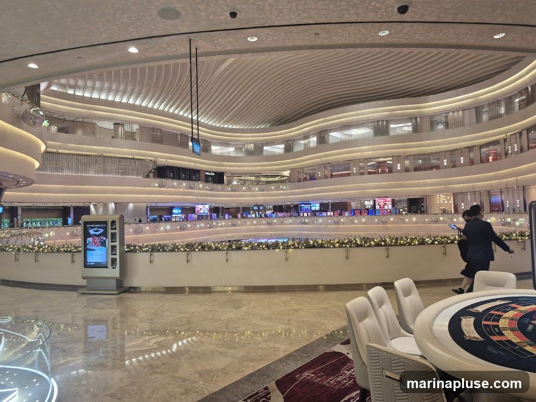 A wide shot of a luxurious casino interior with multiple levels. The architecture features curved ceilings and balconies adorned with festive lights. In the foreground, a roulette table with chairs is visible. In the mid-ground, a digital display kiosk stands next to a railing overlooking the lower level, which is filled with slot machines and gaming areas. People can be seen walking around the various levels.